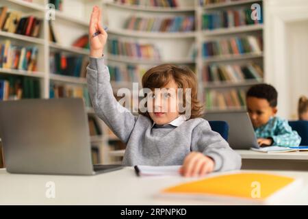 Scolaro intelligente che alza la mano per rispondere a una domanda in classe di alfabetizzazione digitale, seduto di fronte a un notebook Foto Stock