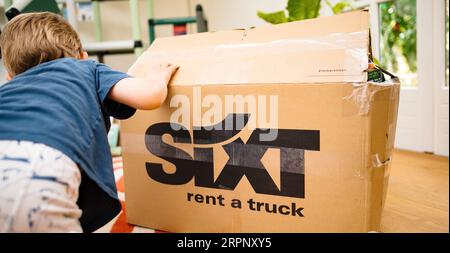 Brema, Germania - 14 agosto 2023: Una mano per bambini sulla scatola in un soggiorno, che indica sixt, affittare scatole di cartone per camion. Si stanno preparando a trasferirsi in una nuova casa o appartamento. La vista è da dietro il bambino. Foto Stock