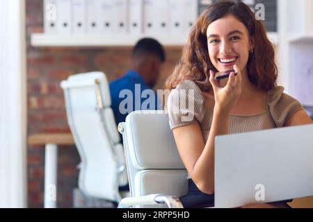 Ritratto di una giovane donna d'affari sorridente che lavora alla scrivania in ufficio e parla con il microfono del telefono cellulare Foto Stock
