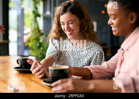 Due giovani amiche che si incontrano in un bar e guardano insieme il telefono cellulare Foto Stock