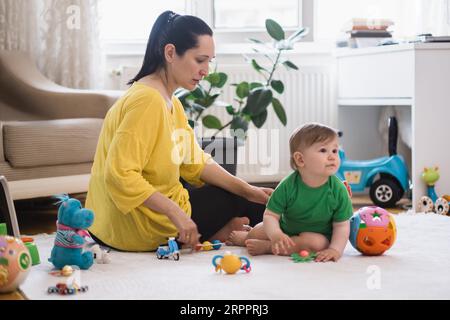 Una madre amorevole che mostra giocattoli diversi a un bambino seduto sul tappeto bianco sul pavimento Foto Stock