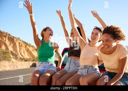 Le amiche che ridono sedute sul cofano di un'auto a tetto aperto durante un viaggio in auto Foto Stock