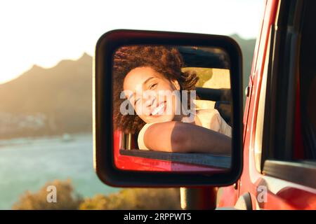 Ritratto di una donna che ride riflessa nello specchio dell'ala dell'auto che si diverte in un'auto a tetto aperto in viaggio su strada Foto Stock