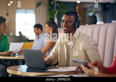 Uomo d'affari che indossa cuffie wireless che lavora su un computer portatile in un'area salotto informale dell'ufficio moderno Foto Stock