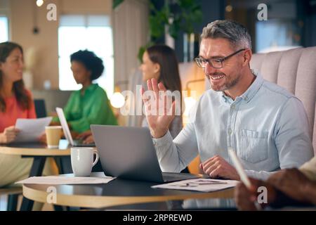 Un uomo d'affari maturo che effettua videochiamate sul computer portatile nell'area salotto informale dell'ufficio moderno Foto Stock
