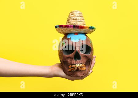 Donna che tiene a mano il cranio umano dipinto per il giorno dei morti del Messico (El dia de Muertos) con sombrero su sfondo giallo Foto Stock