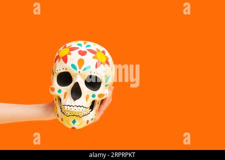 Mano femminile che tiene il cranio umano dipinto per il giorno dei morti del Messico (El dia de Muertos) su sfondo arancione Foto Stock