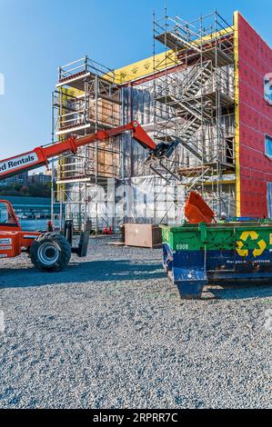 Un ascensore a noleggio arancione con braccio esteso fino al tetto dell'edificio in un cantiere, mentre un operaio edile in un elmetto cammina sul posto. Foto Stock