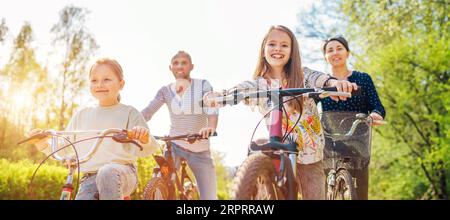 Sorridente padre e madre con due figlie durante l'estate in bicicletta all'aperto. Amano stare insieme nel parco estivo della città. Felice paternità a Foto Stock