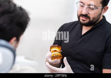 Medico serio con occhiali che mostrano mascelle umane modello con supporti dentali al paziente in clinica Foto Stock