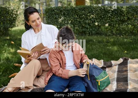 ragazzo con sindrome di down che prende il libro dallo zaino scolastico vicino a madre felice su coperta nel parco Foto Stock