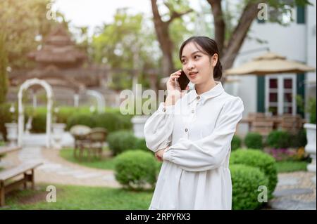 Una bella giovane donna asiatica sta parlando al telefono con qualcuno mentre si rilassa in un bellissimo giardino o cortile. Concetto di persone e tecnologia Foto Stock