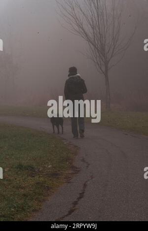 Un maschio adulto che cammina il suo cane domestico su un marciapiede in un parco avvolto dalla nebbia Foto Stock