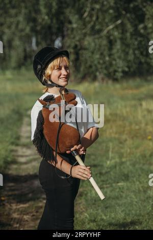 Ritratto di una ragazza - un fantino in un casco e una maglietta bianca, che abbraccia un cavallo marrone - un giocattolo su un bastone, guarda nella macchina fotografica. Alberi verdi nel ba Foto Stock