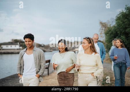 Giovani amici maschi e femmine che bevono mentre camminano insieme vicino al lago Foto Stock