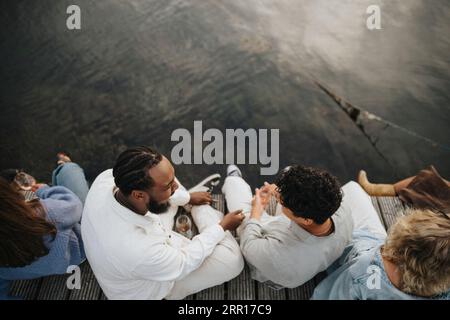 Vista ad alto angolo di amici maschi e femmine che si divertono sedendo sul molo vicino al lago Foto Stock