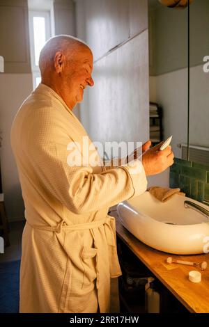 Uomo anziano sorridente mentre si applica l'idratante in bagno a casa Foto Stock