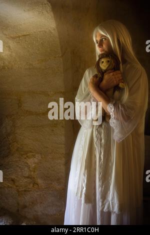 Fantasma femminile che indossa un antico abito da notte bianco con in mano una bambola raccapricciante Foto Stock