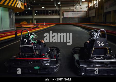 due diversi piloti con caschi e abbigliamento sportivo che guidano go-kart sul circuito indoor, concorrenti Foto Stock