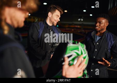 allegri uomini multiculturali che chiacchierano all'interno del circuito di kart, tre piloti di kart che reggono caschi Foto Stock