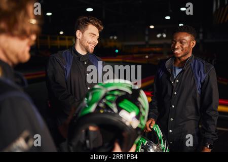 felici uomini multiculturali che chiacchierano all'interno del circuito di kart, tre piloti di kart che reggono elmetti Foto Stock