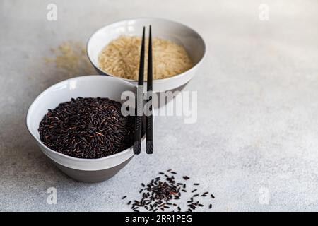 Angolo elevato di riso nero crudo selvatico e riso bianco sbucciato nelle ciotole con bacchette contro la superficie bianca Foto Stock