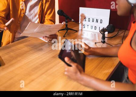 Dall'alto ritagliate una femmina nera con le cuffie per tablet che guardano il maschio anonimo con gesti della mano mentre state seduti a registrare podcast con i microfoni Foto Stock