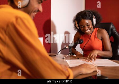 Uomo ispanico sorridente che guarda lontano e donna nera felice che guarda i fogli musicali con microfoni e registra podcast in remoto Foto Stock