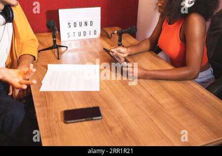 Dall'alto ritagliate una femmina nera con le cuffie per tablet che guardano il maschio anonimo con gesti della mano mentre state seduti a registrare podcast con i microfoni Foto Stock
