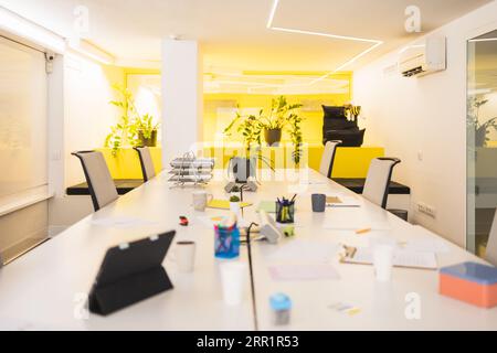 Sala riunioni con scrivania, tavolo lungo con sedie per tablet, piante in vaso su una scrivania decorata con pareti in vetro giallo e corridoio esterno sfocato Foto Stock