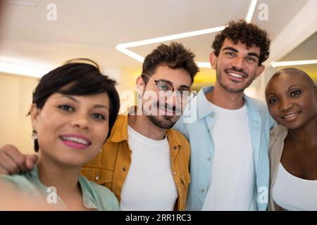 Colleghi positivi e diversi che sorridono e guardano la fotocamera mentre sono in piedi vicino alle luci dell'ufficio e scattano selfie insieme Foto Stock