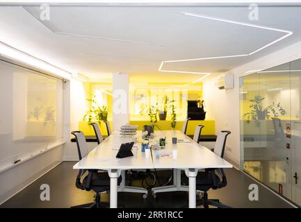 Sala riunioni con scrivania, tavolo lungo con sedie per tablet, piante in vaso su una scrivania decorata con pareti in vetro giallo e corridoio esterno sfocato Foto Stock