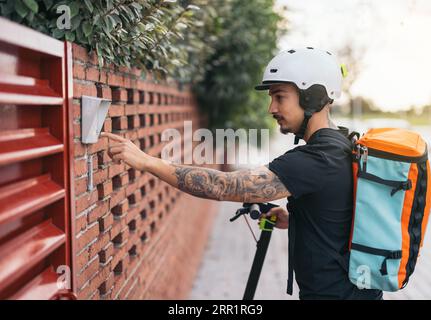 Vista laterale del giovane uomo con casco e zaino termico su uno scooter elettrico che suona il campanello della casa Foto Stock