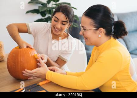 Amiche felici che guardano in basso mentre si siedono sedute a un tavolo di legno con attrezzi di Halloween e occhi intagliati alla luce del giorno Foto Stock