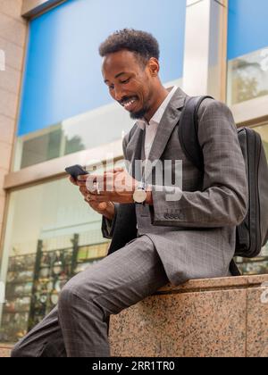 Un ragazzo afro-americano con abiti formali seduto su una panchina di pietra e un cellulare alla luce del sole Foto Stock