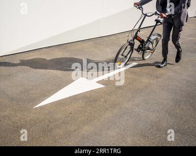 Vista laterale di una persona anonima corta in bicicletta lungo la strada asfaltata con puntatore vicino alla parete bianca Foto Stock