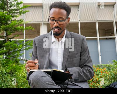 Uomo afro-americano focalizzato con occhiali in abito formale seduto sulla strada della città e scrivendo appunti su un quaderno Foto Stock