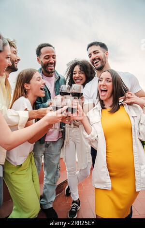 Verticale. Gruppo di giovani amici allegri sorridendo e brindando bicchieri di vino rosso alla festa di compleanno sul tetto. Felici adulti che si divertono a indossare occhiali da vista durante la festa del fine settimana. Amici con alcol. Foto di alta qualità Foto Stock