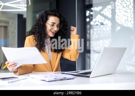 Donna d'affari di successo che fa i documenti, capo con gli occhiali seduti alla scrivania sul posto di lavoro utilizzando un computer portatile che lavora con i documenti, che tiene la mano per celebrare la vittoria e il trionfo. Foto Stock