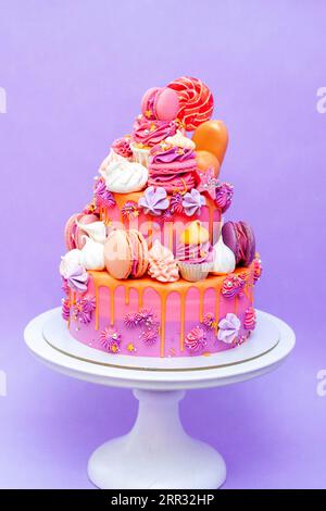 Torta sfumata rosa, viola e arancia con rose al formaggio spalmabile, macaron, cupcake, meringhe su fondo liscio viola Foto Stock