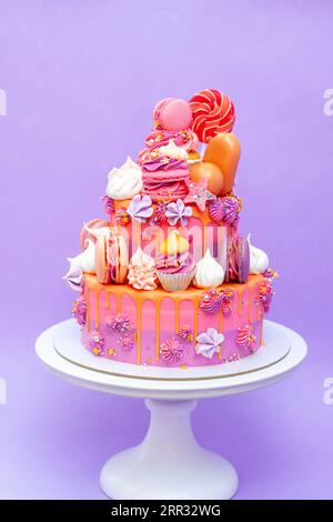 Torta sfumata rosa, viola e arancia con rose al formaggio spalmabile, macaron, cupcake, meringhe su fondo liscio viola Foto Stock