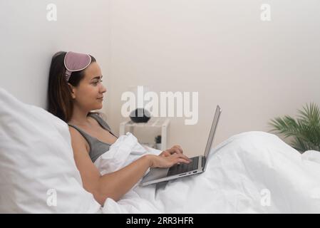 Una giovane donna sorridente scrive messaggi agli amici sul computer portatile seduti a letto in una camera da letto accogliente la mattina Foto Stock