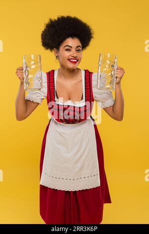 felice cameriera afroamericana dell'oktoberfest con un abito in stile dirndl che tiene le tazze di birra vuote sul giallo Foto Stock