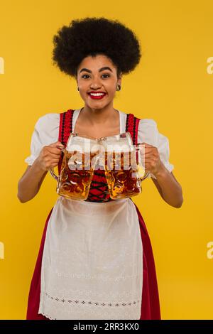 allegra cameriera afroamericana in abbigliamento oktoberfest con tazze da birra e sorridendo sul giallo Foto Stock