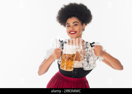 una cameriera bavarese afro-americana con tazze di birra piene e vuote su bianco, oktoberfest Foto Stock