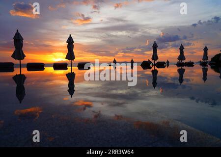 Ombrelloni chiusi che si riflettono in una piscina a sfioro affacciata sul mare in un suggestivo cielo al tramonto Foto Stock