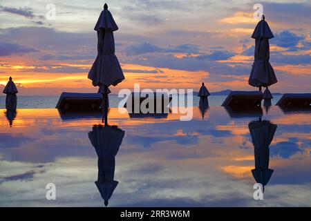 Ombrelloni chiusi che si riflettono in una piscina a sfioro affacciata sul mare in un suggestivo cielo al tramonto Foto Stock