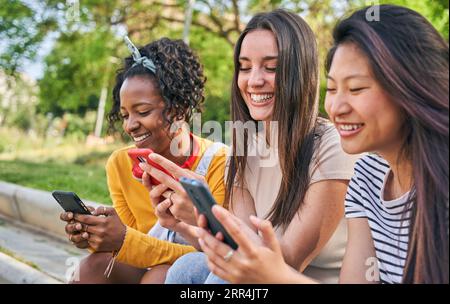 Tre giovani donne sorridenti che usano il cellulare all'aperto. Gruppo di amici felici che controllano i social network Foto Stock