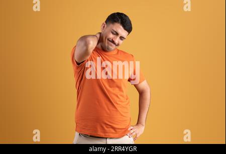 Triste uomo brasiliano in t-shirt che spinge la mano al collo e soffre di dolori muscolari, posando su sfondo giallo Foto Stock