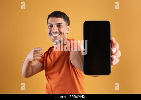 Felice uomo brasiliano che punta in modo informale verso uno smartphone moderno con schermo vuoto, offerta pubblicitaria mobile o sito Web Foto Stock